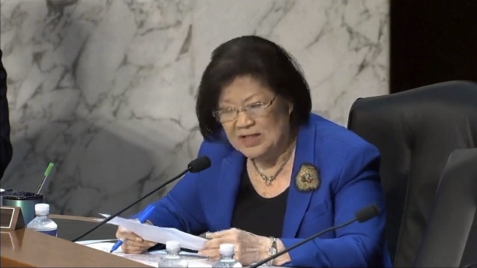 Senator in a blue blazer speaking at a congressional hearing, holding documents, with a marble background and water bottles on the table.