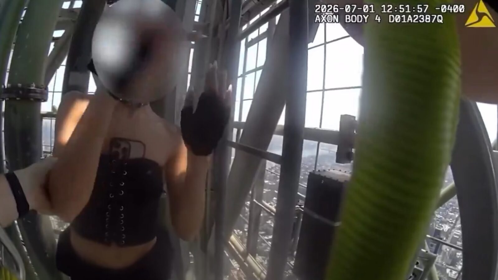 Person in a black corset waving while standing on a high structure with city views in the background, captured from a body camera perspective.