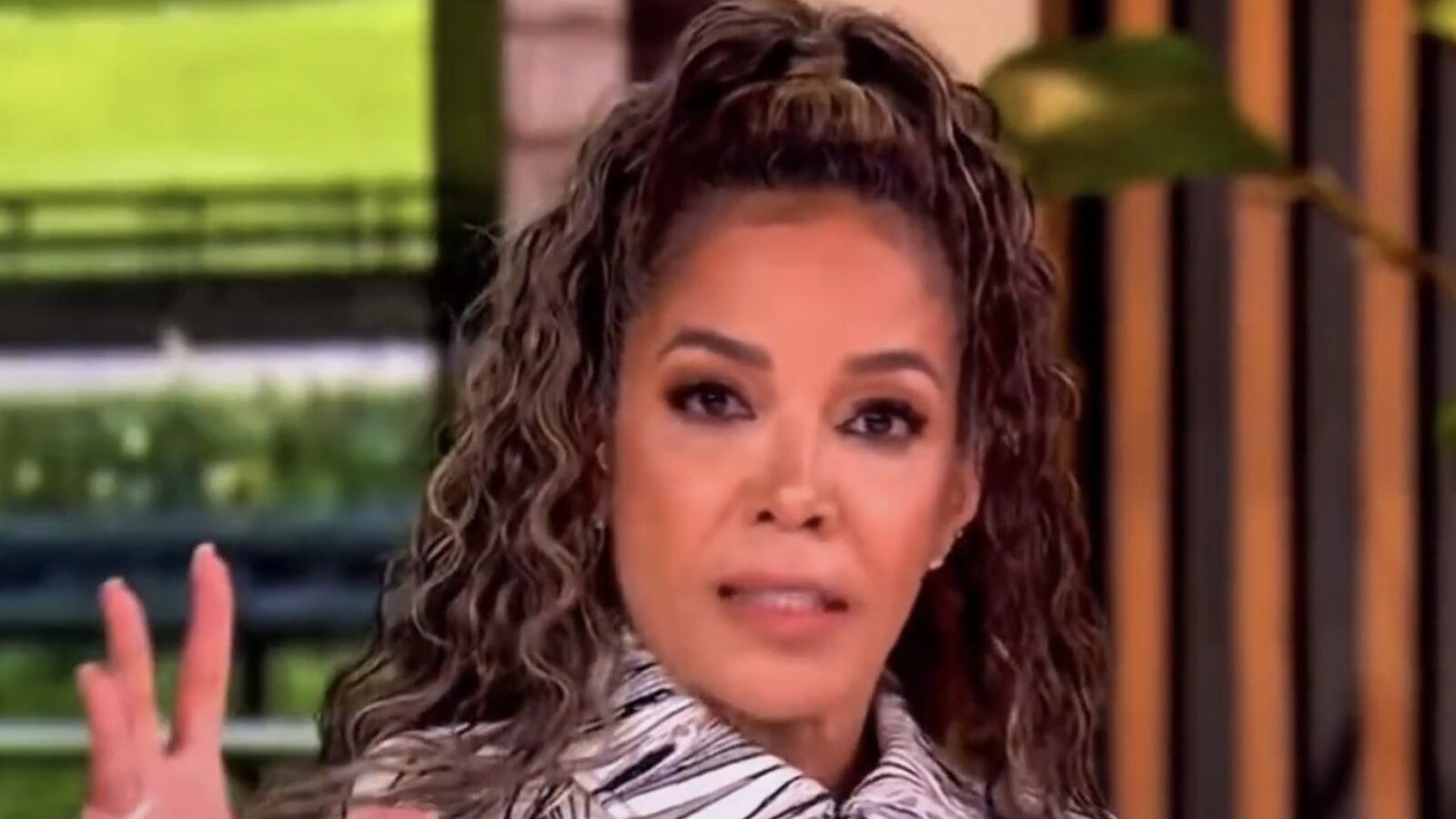 Television host speaking passionately with raised hands, wearing a patterned coat, set against a vibrant background.