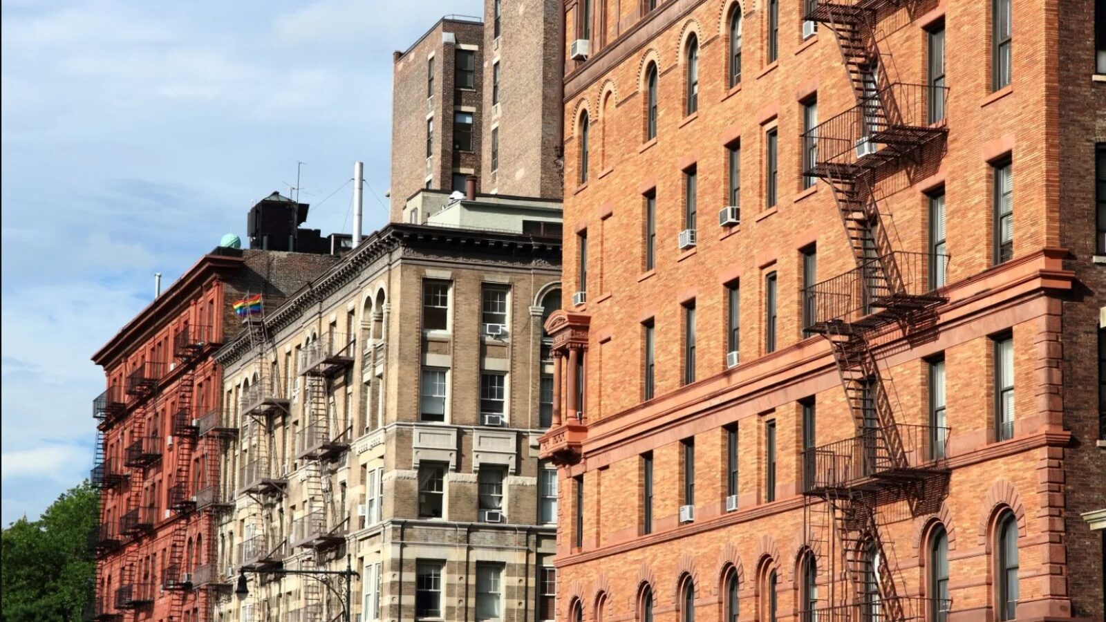 Historic buildings with fire escapes and diverse architectural styles in an urban setting under a partly cloudy sky.