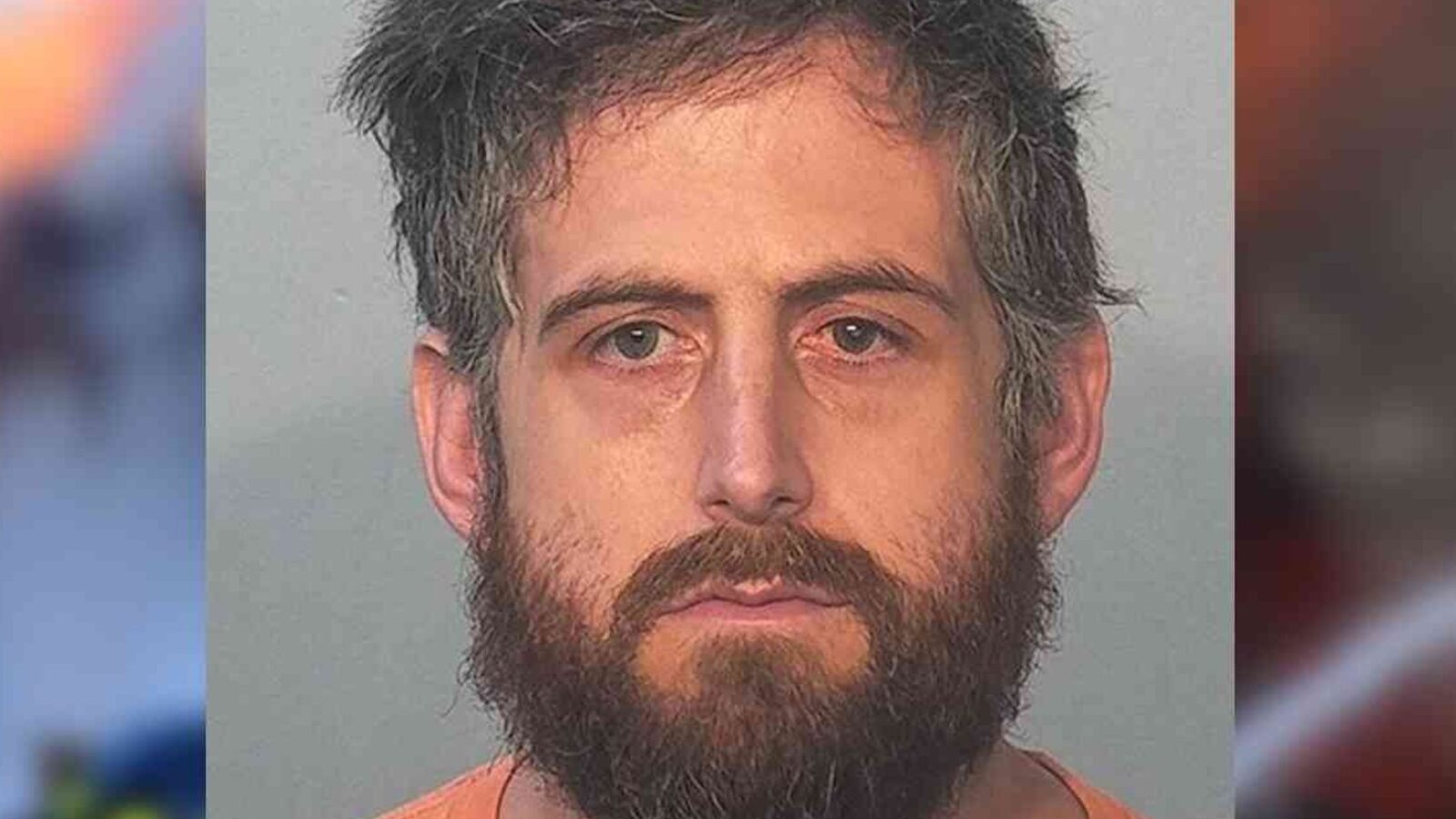 Mugshot of a man with a beard and disheveled hair, wearing an orange shirt, against a neutral background.