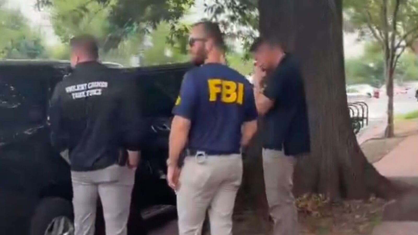 FBI agents in tactical gear discussing near a black vehicle in an urban setting, surrounded by trees and pedestrians.