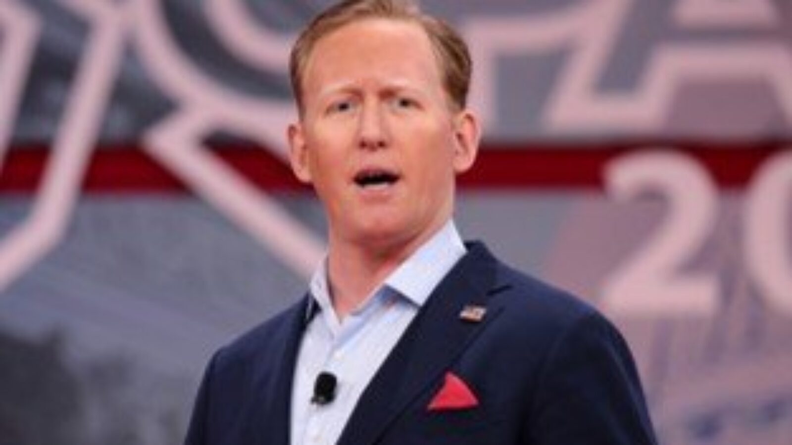 Speaker presenting at a political conference, wearing a dark suit and red pocket square, with a backdrop featuring the event's branding.