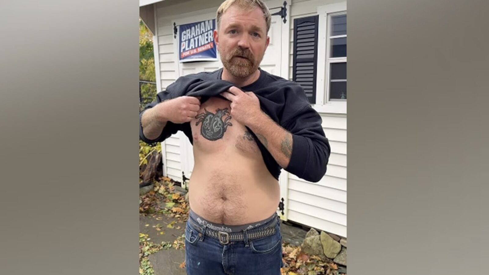 Man lifting his shirt to reveal a tattoo on his chest, standing outside a house with autumn leaves scattered on the ground.