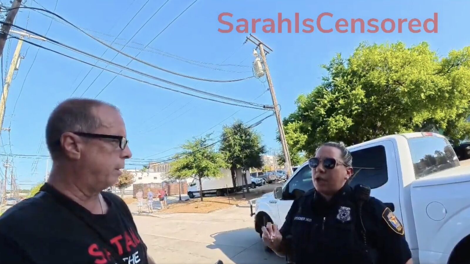 Man in glasses converses with police officer in uniform on a sunny street with power lines and trees in the background.